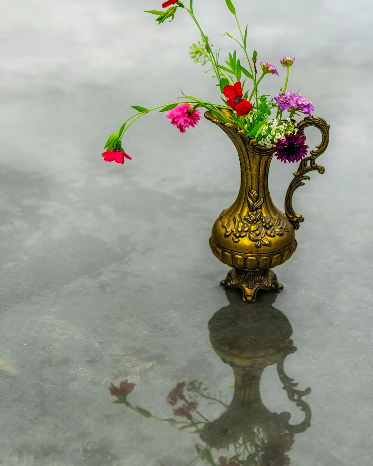 A brass pitcher with wildflowers reflected on a stone surface