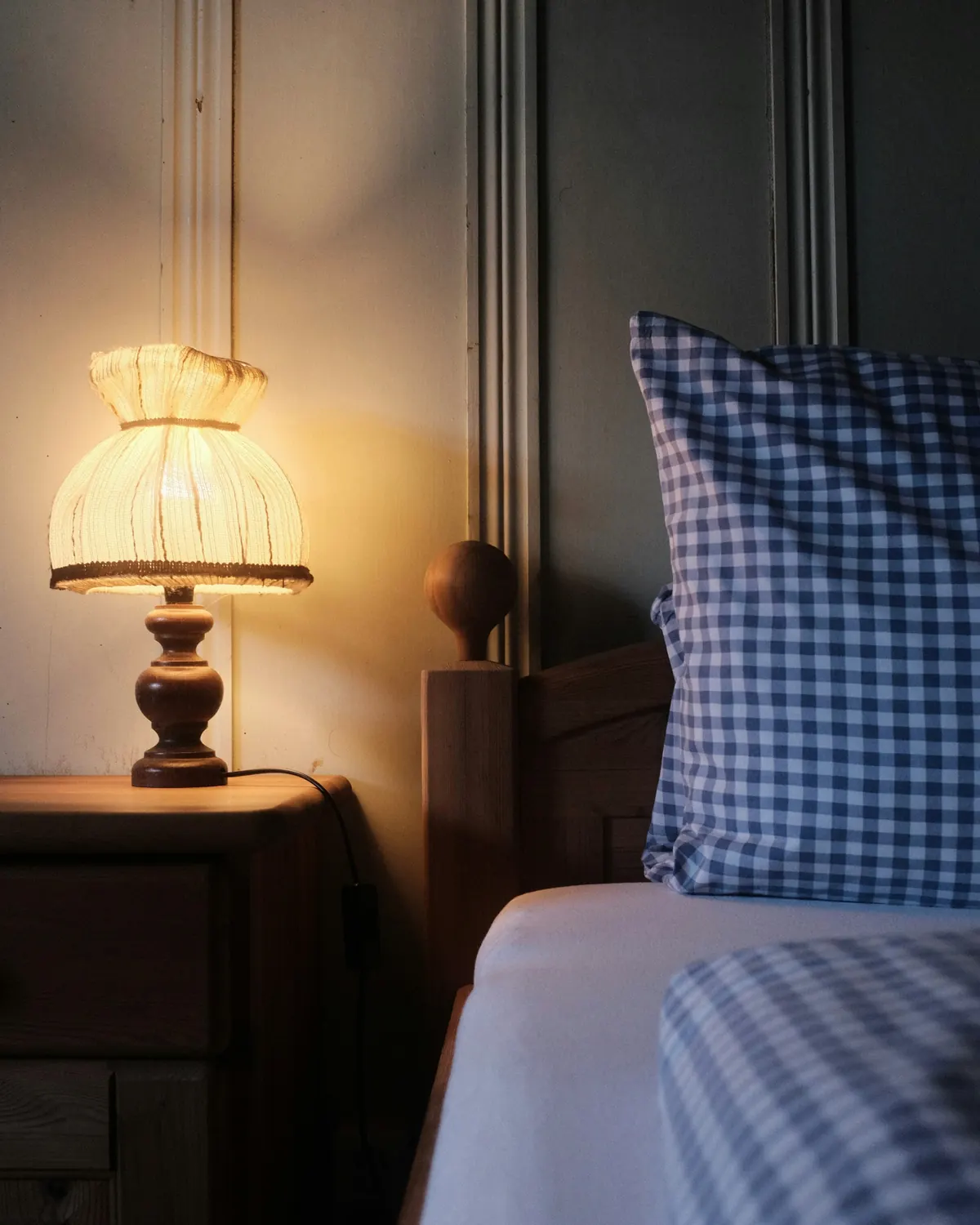A pleated bedside lamp glowing beside gingham bedding