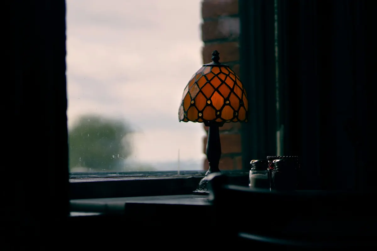 A stained-glass table lamp glowing by a window at dusk