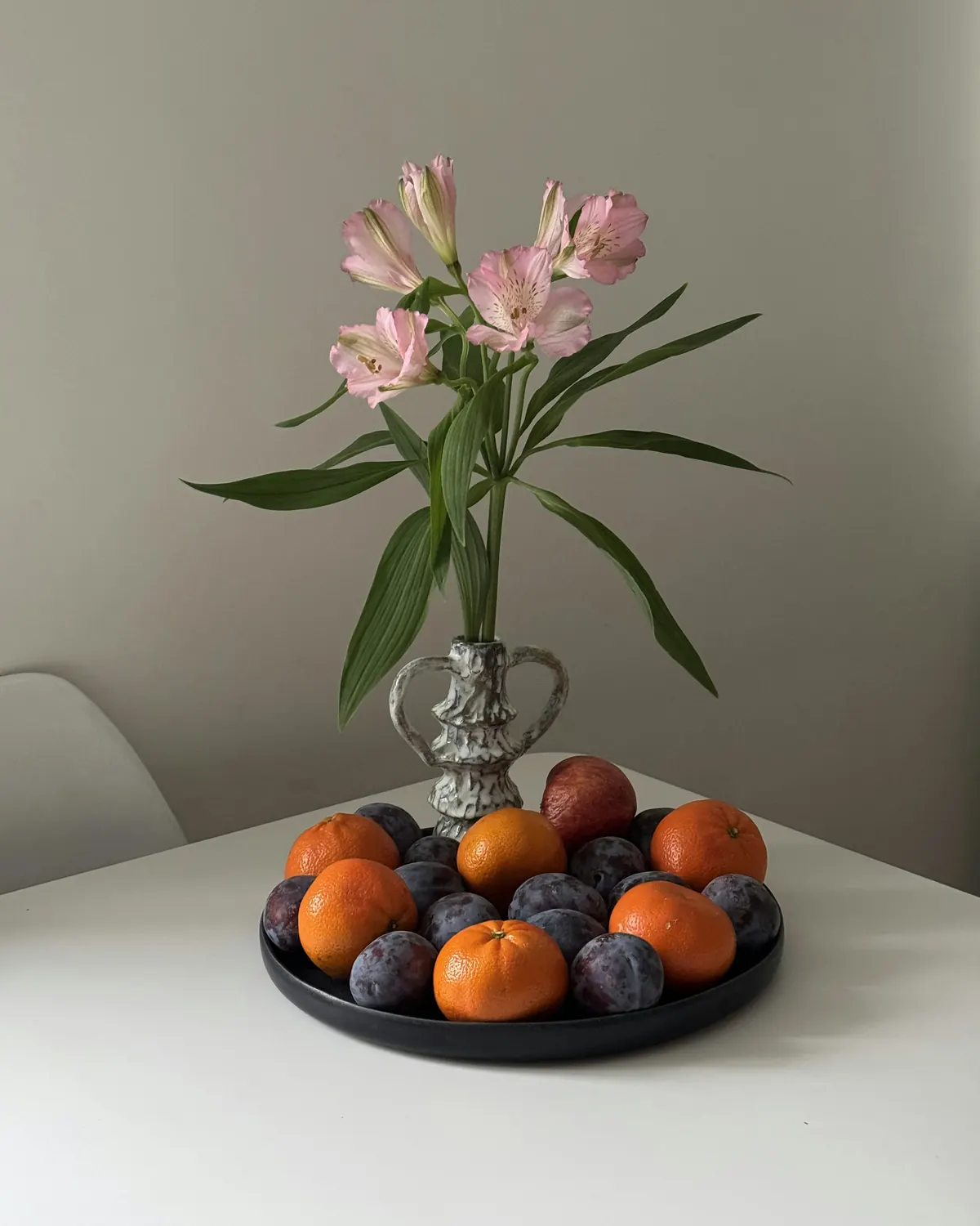 A low dish of mandarins and plums beside a small vase of flowers